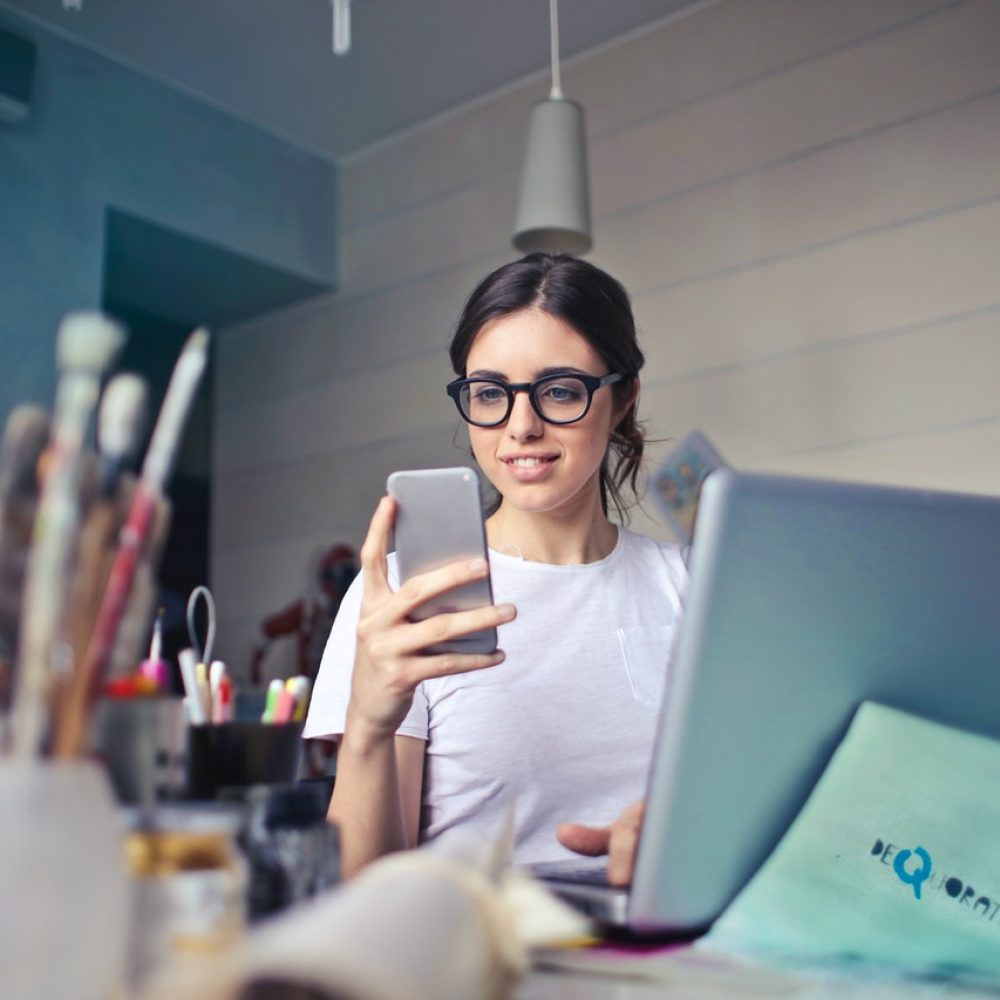 Zadowolona młoda kobieta korzystająca z internetu WiFi 6 w telefonie oraz z laptopa podłączonego do szybkiego internetu światłowodowego w technologii FTTH z prędkością do 10Gbps